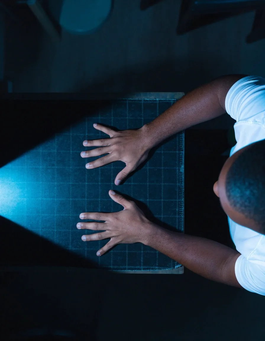 Mãos em postura calma sobre a mesa escura, iluminadas por uma luz azulada vinda de fora do quadro